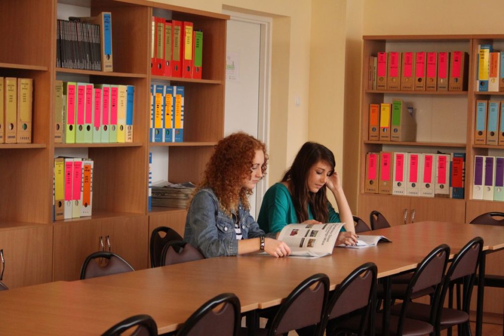 Wnętrze biblioteki szkolnej - drewniane regały z książkami. Przy stole siedzi dwójka uczennic czytających czasopismo  