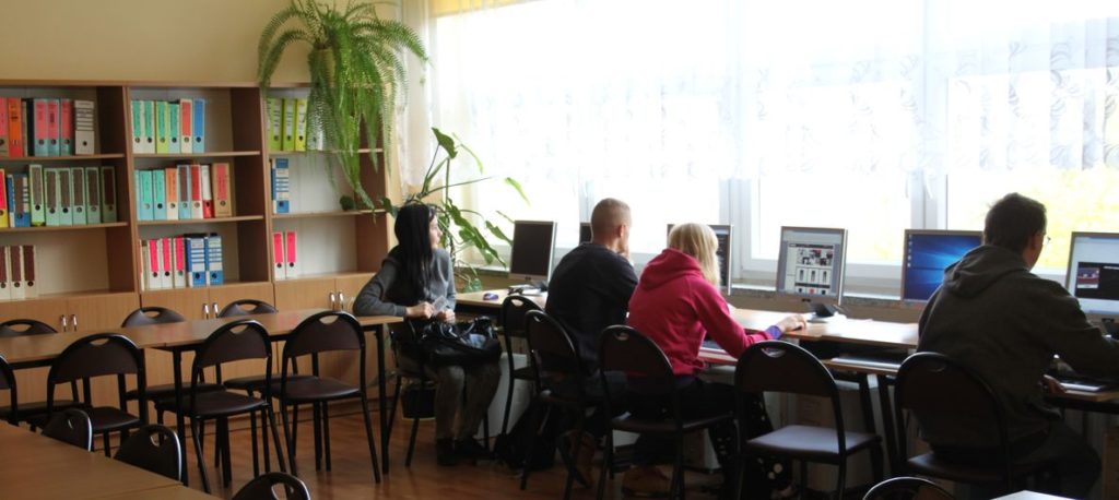 Wnętrze biblioteki szkolnej - drewniane regały z książkami. 4 Uczniów siedzi przy biurku i pracuje przy komputerze.  