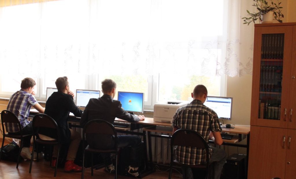 Wnętrze biblioteki szkolnej - drewniane regały z książkami. 4 Uczniów siedzi przy biurku i pracuje przy komputerze.  