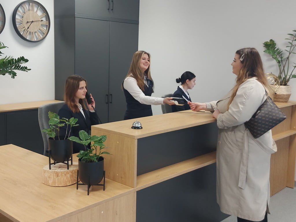 Uczniowie stojący przy recepcji. Jedna uczennica daje klucze do pokoju gościowi hotelowemu