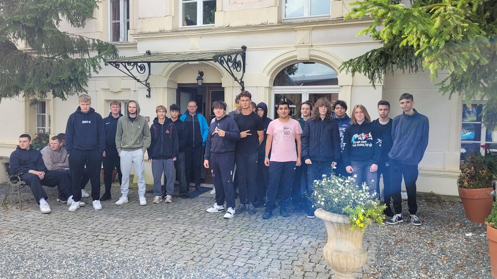 Grupa osób stojąca przed zabytkowym budynkiem z jasną elewacją i ozdobnym wejściem. Po bokach drzwi znajdują się donice z roślinami, a na pierwszym planie duża donica z kwiatami. Twarze osób są niewidoczne.