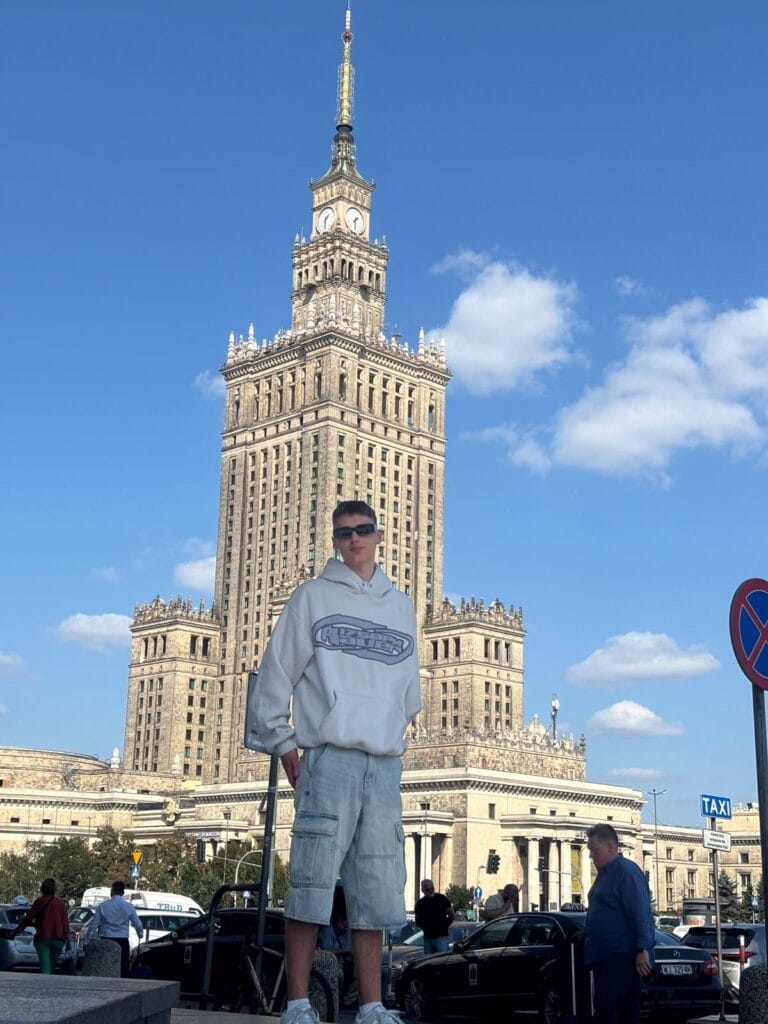 Osoba w białej bluzie i jeansowych spodenkach stoi przed Pałacem Kultury i Nauki w Warszawie, w tle widoczni są przechodnie, samochody oraz znak TAXI