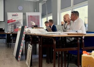 Grupa osób siedząca przy stole konferencyjnym, na którym znajdują się dokumenty, laptopy i materiały promocyjne. W tle widoczne są banery z logo 'URBEX' oraz 'BOLIX', sugerujące tematykę związaną z architekturą lub budownictwem