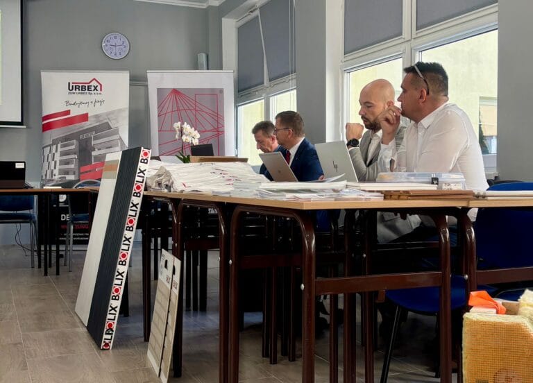 Grupa osób siedząca przy stole konferencyjnym, na którym znajdują się dokumenty, laptopy i materiały promocyjne. W tle widoczne są banery z logo 'URBEX' oraz 'BOLIX', sugerujące tematykę związaną z architekturą lub budownictwem