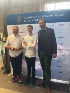 Trzech mężczyzn stoi przed ścianką z logotypami i napisem ‘AKADEMIA MISTRZÓW SMAKU BY BELVEDERE’. Dwaj z nich ubrani są w białe kucharskie bluzy i czarne spodnie, trzeci ma na sobie ciemną marynarkę, niebieską koszulę, zielone spodnie i brązowe buty