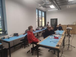 Uczniowie podczas zajęć technologicznych w Inkubatorze Przedsiębiorczości. Trzy osoby pracują przy stole z niebieskimi matami, narzędziami i komponentami elektronicznymi. Jedna osoba siedzi osobno, obserwując grupę. W tle widoczne są duże okna, półki z wyposażeniem i sprzęt edukacyjny. Zdjęcie przedstawia atmosferę praktycznej nauki i współpracy