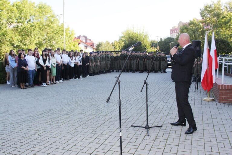 Mężczyzna przemawia do mikrofonu podczas uroczystości na zewnątrz; wokół niego osoby w strojach formalnych i mundurach, na pierwszym planie dwa mikrofony na statywach, po prawej stronie dwie flagi, w tle drzewa i budynki