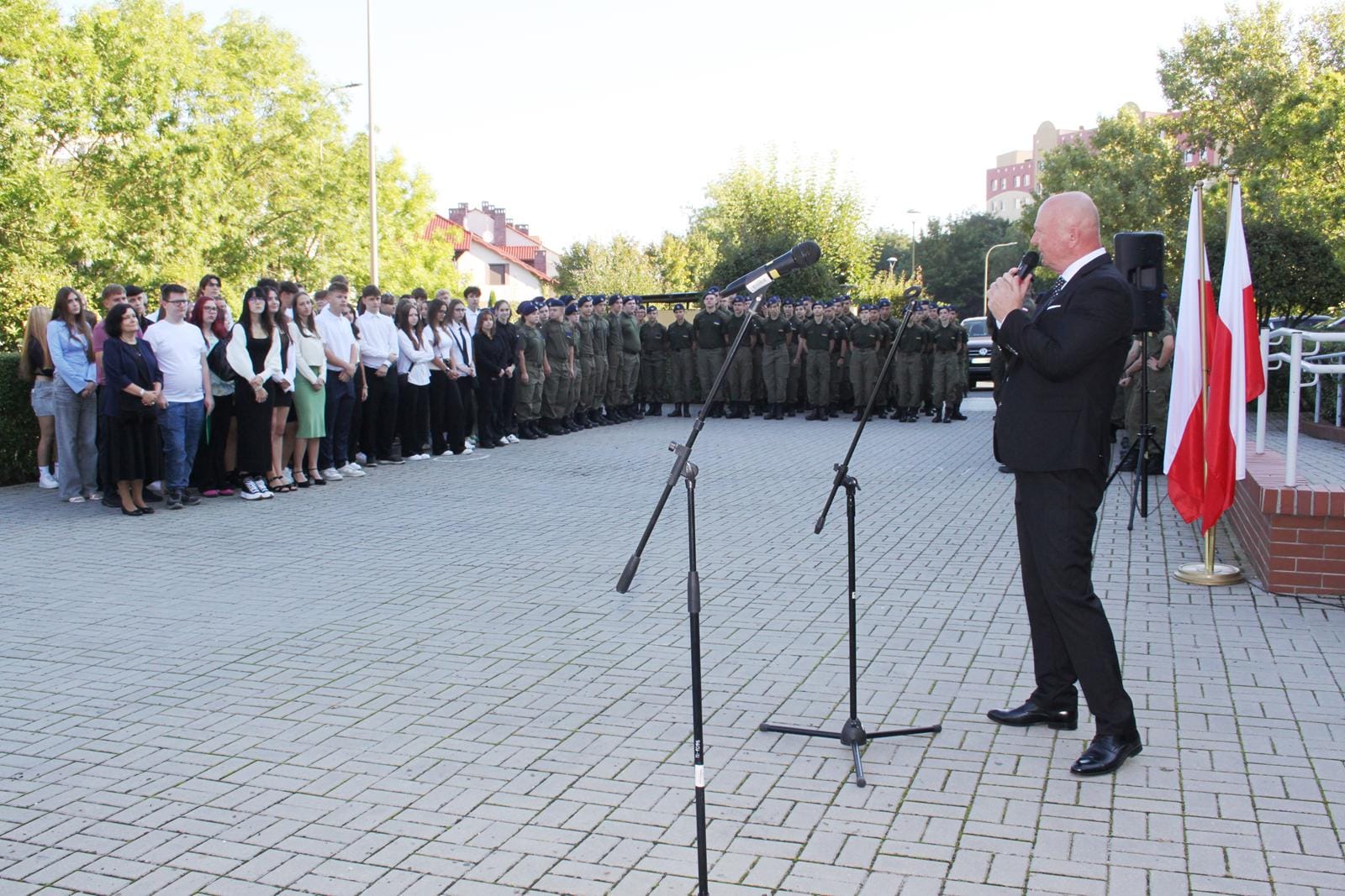 Mężczyzna przemawia do mikrofonu podczas uroczystości na zewnątrz; wokół niego osoby w strojach formalnych i mundurach, na pierwszym planie dwa mikrofony na statywach, po prawej stronie dwie flagi, w tle drzewa i budynki