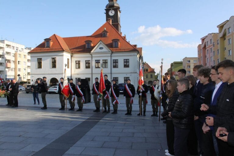 Grupa osób w mundurach z flagami stoi przed zabytkowym budynkiem z wieżą zegarową i czerwonym dachem, prawdopodobnie podczas oficjalnej uroczystości na rynku miejskim. Po prawej stronie widoczna jest publiczność obserwująca wydarzenie.