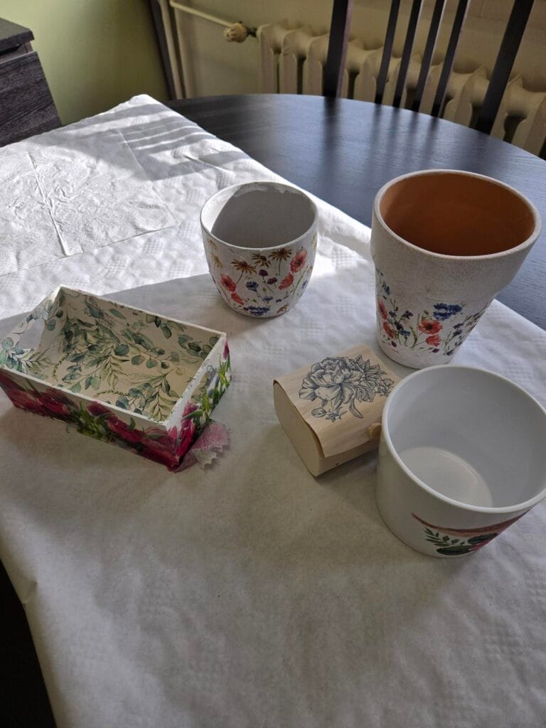Na stole leżą ozdobione przedmioty wykonane techniką decoupage: dwie doniczki z kolorowymi kwiatowymi wzorami, biała skrzynka z motywem roślinnym oraz małe drewniane pudełko z niebieskim kwiatowym ornamentem