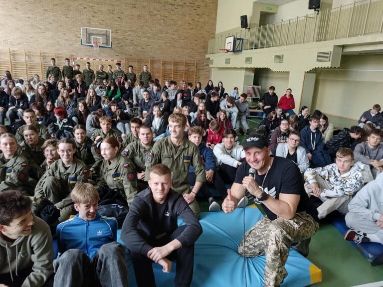 Grupa uczniów siedzi w sali gimnastycznej, część w strojach wojskowych, słuchając osoby prowadzącej z mikrofonem. W tle widoczne są drabinki gimnastyczne, kosz do koszykówki oraz balkon z barierkami