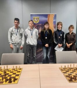 Pięć osób stoi w sali przy roll-upie z herbem i grafiką, mają na szyjach medale. Na pierwszym planie widać stół z rozstawionymi szachownicami i figurami szachowymi