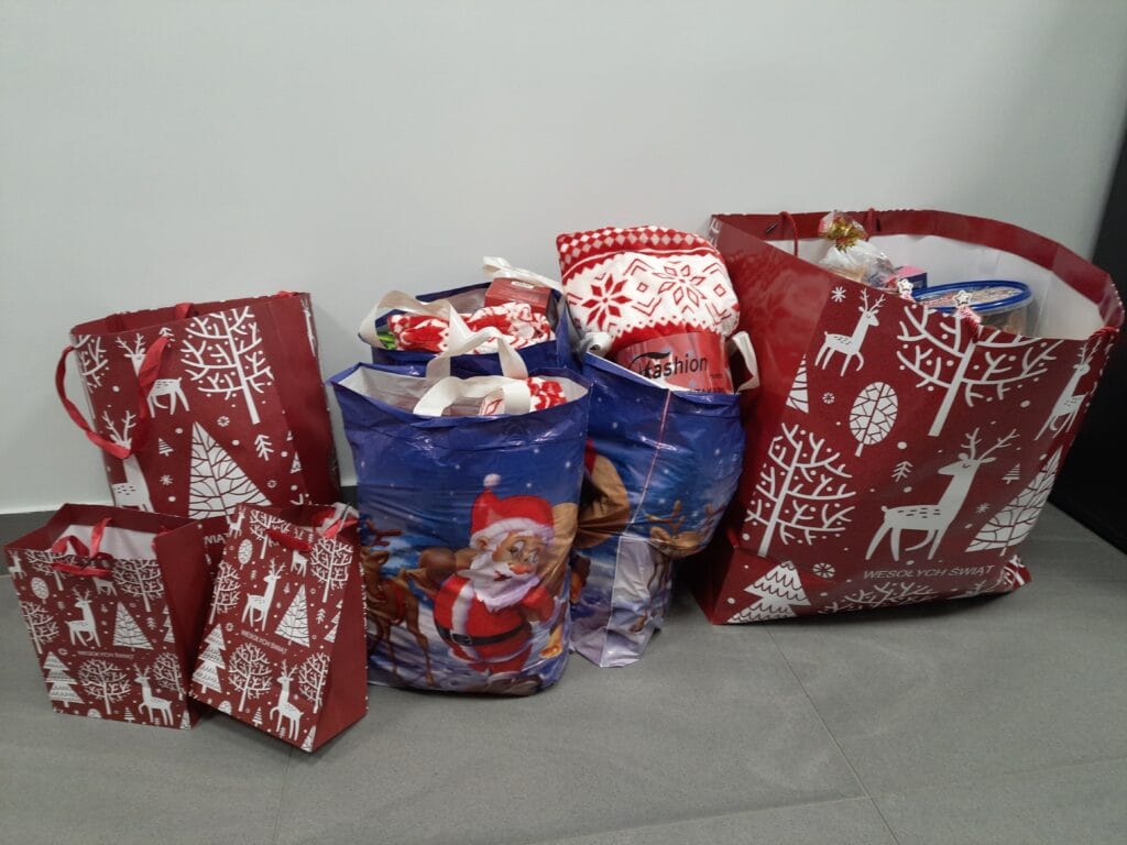 Kilka dużych i małych toreb prezentowych w świątecznych wzorach, wypełnionych artykułami spożywczymi i upominkami, ustawionych na podłodze przy ścianie