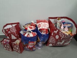 Kilka dużych i małych toreb prezentowych w świątecznych wzorach, wypełnionych artykułami spożywczymi i upominkami, ustawionych na podłodze przy ścianie