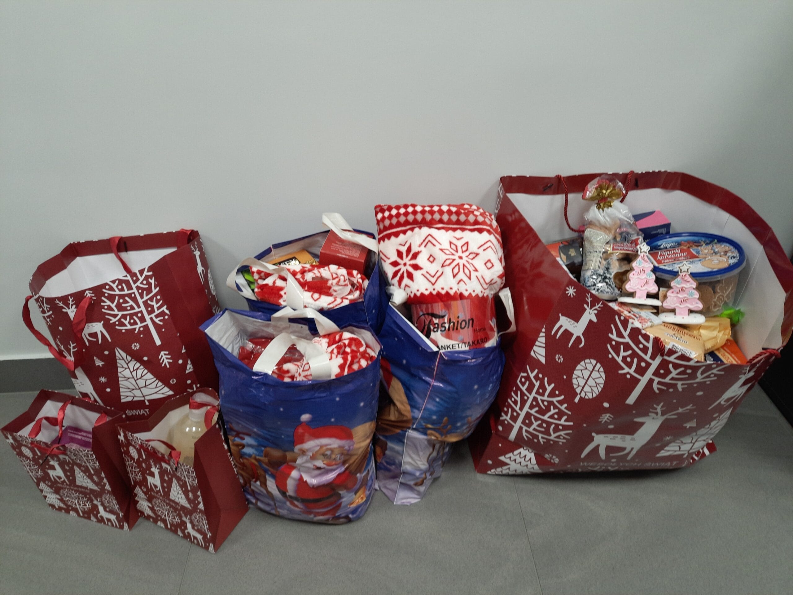 Kilka dużych i małych toreb prezentowych w świątecznych wzorach, wypełnionych artykułami spożywczymi i upominkami, ustawionych na podłodze przy ścianie