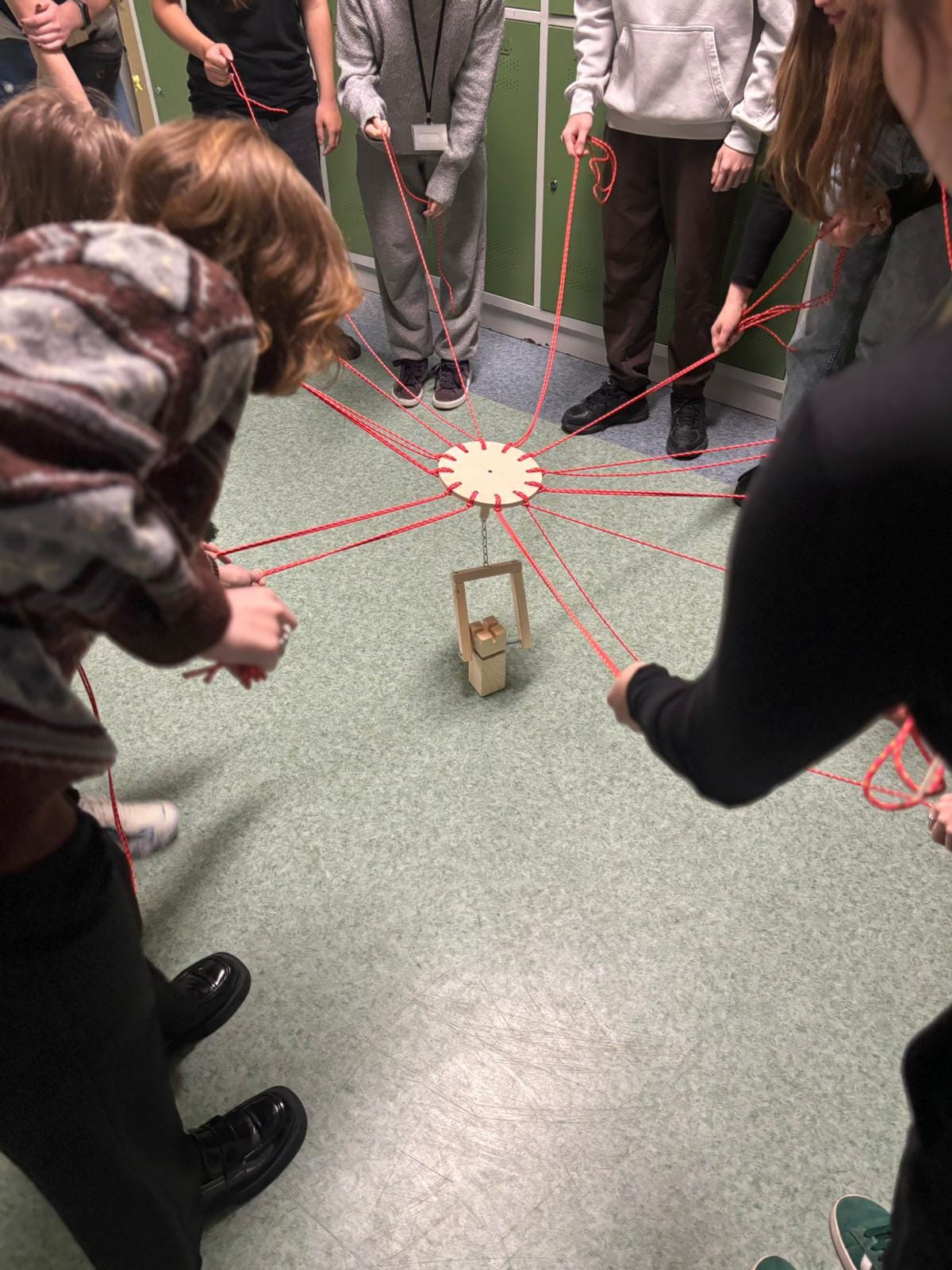 Grupa osób stoi w kręgu, trzymając czerwone sznurki przymocowane do drewnianego elementu w centrum; na podłodze znajduje się mała drewniana konstrukcja, którą wspólnie manipulują