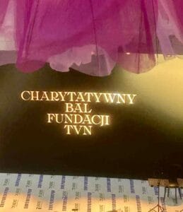 Czarny panel z podświetlonym złotym napisem: ‘Charytatywny Bal Fundacji TVN’. U góry widoczne fragmenty dekoracji z różowego tiulu, na dole podłoga z wzorem w jasnych i ciemnych pasach z napisem ‘TVN’.