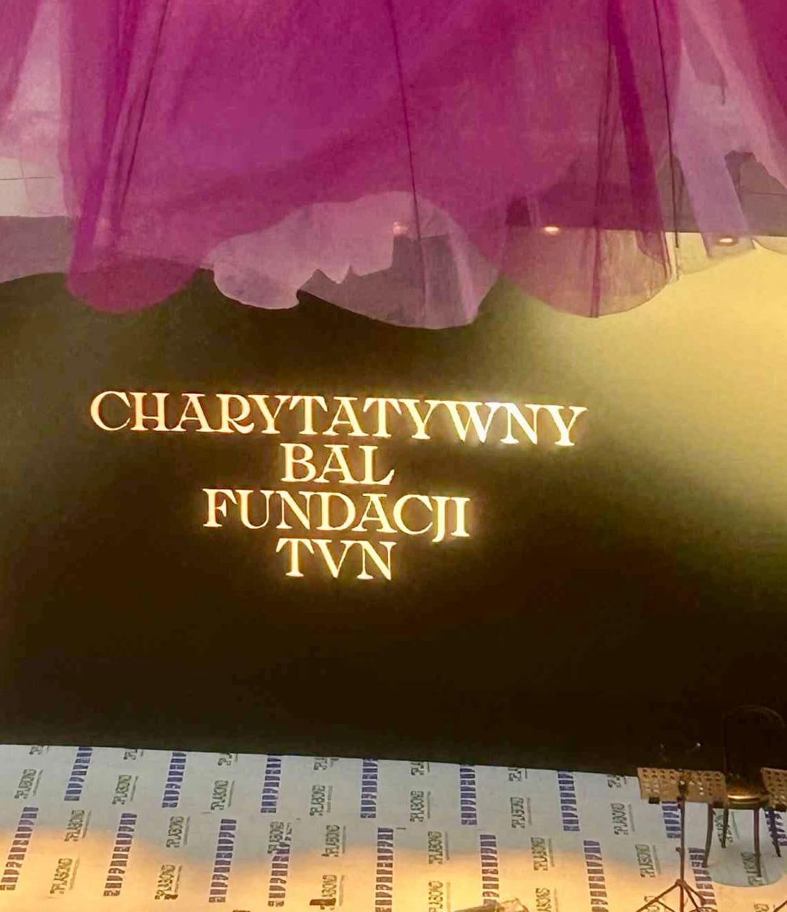 Czarny panel z podświetlonym złotym napisem: ‘Charytatywny Bal Fundacji TVN’. U góry widoczne fragmenty dekoracji z różowego tiulu, na dole podłoga z wzorem w jasnych i ciemnych pasach z napisem ‘TVN’.