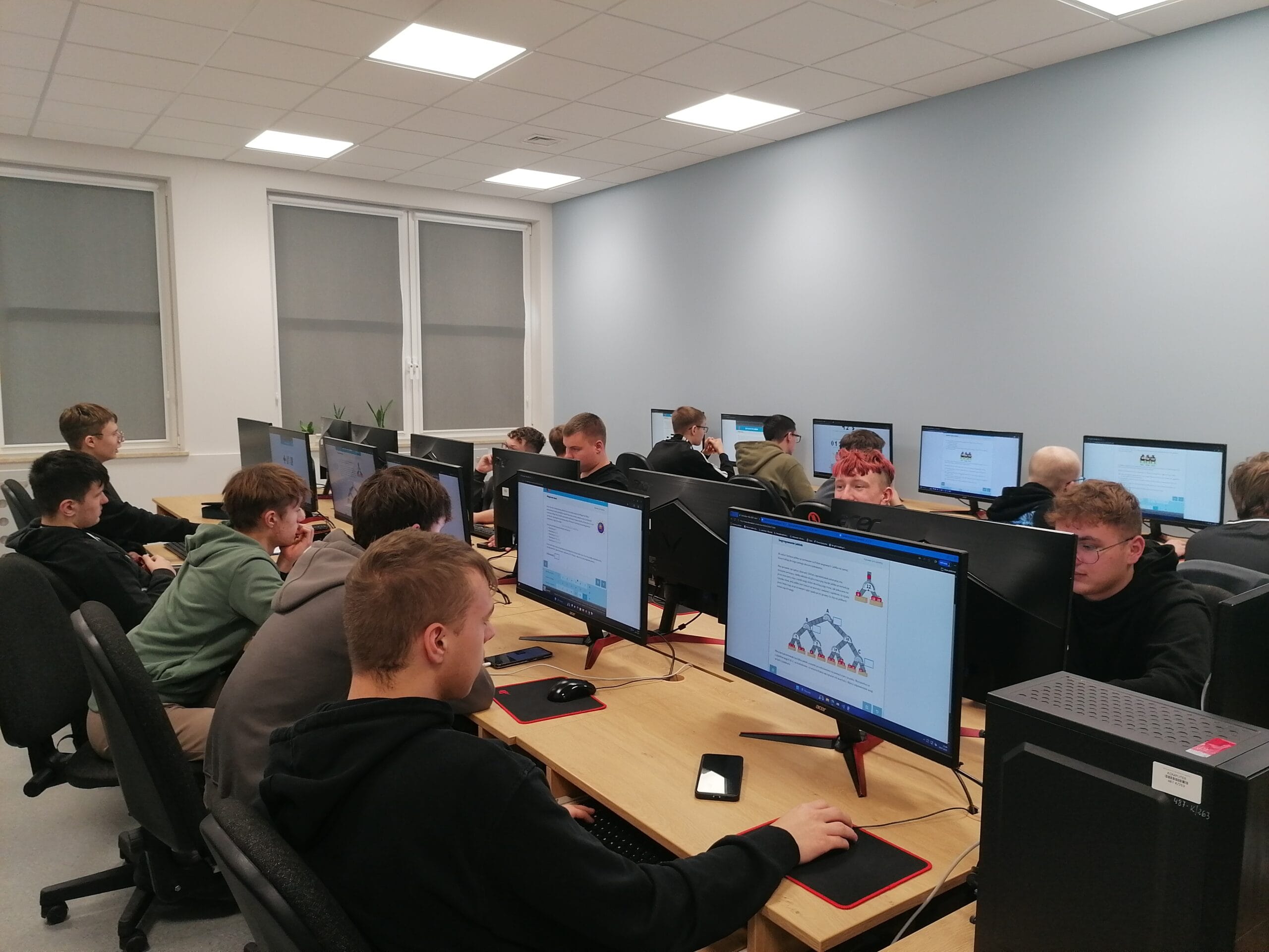 Zdjęcie w sali komputerowej: kilkanaście osób siedzi przy dwóch rzędach biurek z monitorami. Na ekranach widoczne są strony internetowe i schematy drzew binarnych. Na biurkach znajdują się klawiatury, myszki i smartfony. W tle jasne ściany, okna z roletami i sufit z panelami LED