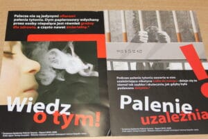 Ulotka antynikotynowa składająca się z dwóch części. Lewa strona przedstawia osobę wydychającą dym papierosowy, z dużym czerwonym napisem ‘Wiedz o tym!’ oraz tekstem ostrzegającym, że bierne palenie jest groźne dla zdrowia i może być śmiertelne. Prawa strona pokazuje dłonie trzymające pręty krat, z napisem ‘Palenie uzależnia!’ i informacją, że nikotyna szybko trafia do mózgu, działając jak podanie dożylne
