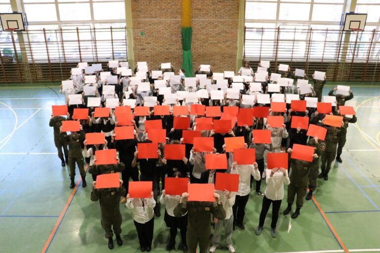 Grupa osób w sali gimnastycznej tworzy flagę Polski, trzymając nad głową białe i czerwone kartony ułożone w poziome pasy