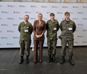 Cztery osoby stoją na tle ścianki z logotypami Ministerstwa Edukacji i Nauki; trzy osoby ubrane w zielone mundury wojskowe, jedna w brązowy garnitur, wszystkie mają identyfikatory