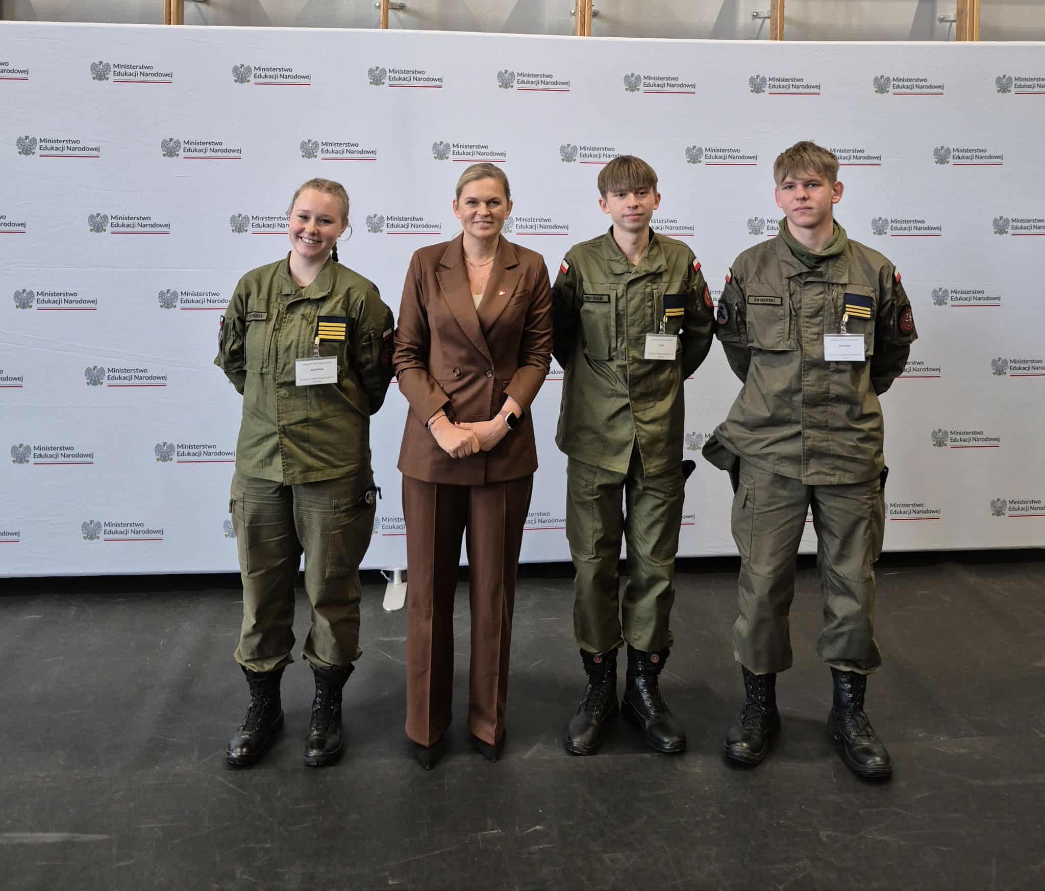 Cztery osoby stoją na tle ścianki z logotypami Ministerstwa Edukacji i Nauki; trzy osoby ubrane w zielone mundury wojskowe, jedna w brązowy garnitur, wszystkie mają identyfikatory