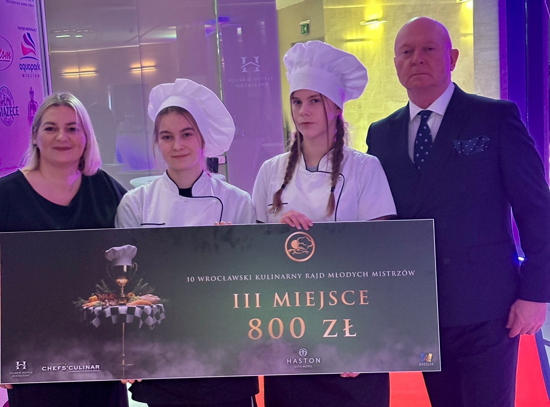 Grupa czterech osób stoi na czerwonym dywanie podczas wydarzenia kulinarnego. Dwie osoby w strojach kucharskich trzymają duży baner z napisem: ‘III miejsce 800 zł’ oraz informacją o 10. Wrocławskim Kulinarnym Rajdzie Młodych Mistrzów. W tle widoczne są logotypy partnerów i dekoracje