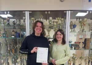 Dwie osoby stoją przed gablotą pełną pucharów i trofeów. Jedna osoba trzyma w dłoniach certyfikat Cambridge English First (FCE), na którym widoczne są szczegóły egzaminu i wyniki