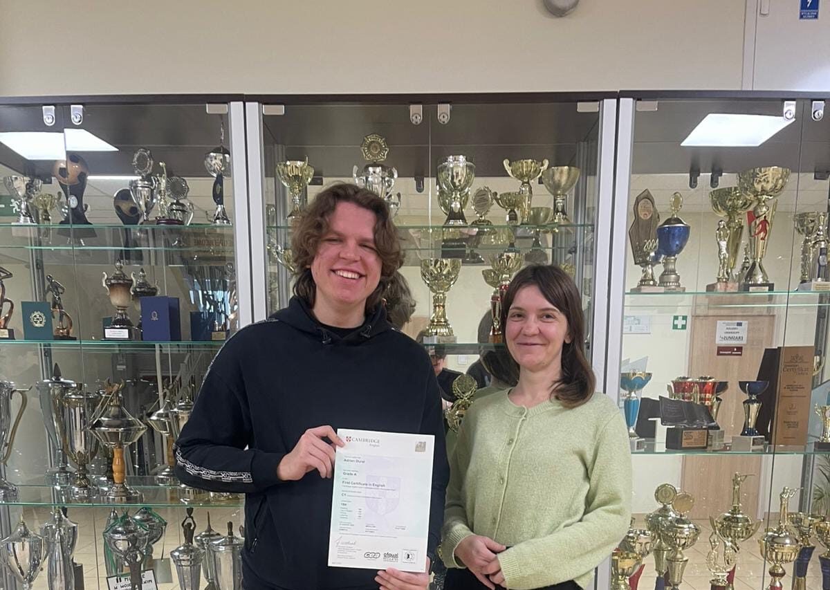 Dwie osoby stoją przed gablotą pełną pucharów i trofeów. Jedna osoba trzyma w dłoniach certyfikat Cambridge English First (FCE), na którym widoczne są szczegóły egzaminu i wyniki
