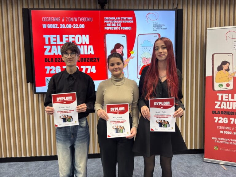 Trzy osoby stojące w pomieszczeniu, każda trzyma dyplom z napisem ‘DYPlOM’ i zdjęciem grupowym. W tle duży ekran z czerwonym napisem ‘Telefon Zaufania dla Dzieci i Młodzieży’ oraz numerem 726 774 774, a obok roll-up z tym samym komunikatem. Na ekranie widoczna grafika przedstawiająca osobę rozmawiającą przez telefon
