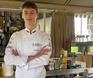 Zdjęcie w przestrzeni kulinarnej: osoba w białej kucharskiej bluzie stoi z założonymi rękami przy stalowym stole roboczym. Na stole znajdują się butelki oleju, przyprawy, rolka ręczników papierowych oraz stos papierowych misek. W tle widoczne są zasłony, duże okna z widokiem na ogród i dekoracyjne elementy sali