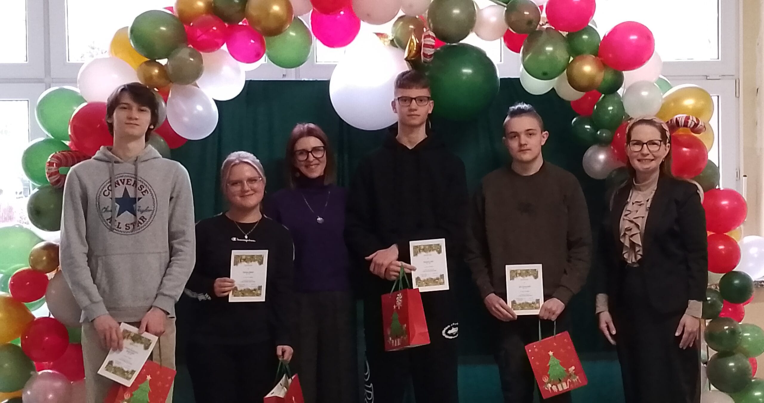 Grupa czterech uczniów i dwóch nauczycielek pozuje do pamiątkowego zdjęcia na tle girlandy z kolorowych balonów. Uczniowie trzymają w dłoniach dyplomy oraz czerwone torebki prezentowe z motywem choinki.