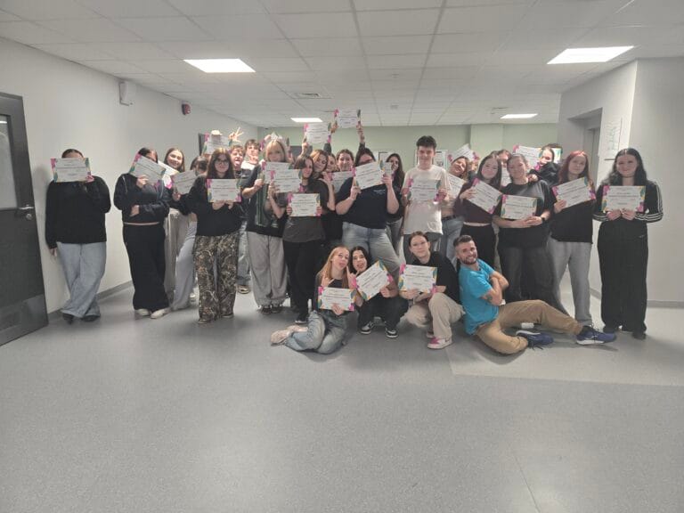 Grupa osób stoi w jasnym pomieszczeniu, trzymając w rękach kolorowe certyfikaty. Uczestnicy ustawieni są w kilku rzędach, część osób siedzi na podłodze z przodu. W tle widoczne są drzwi, jasnoszare ściany i sufit z panelami