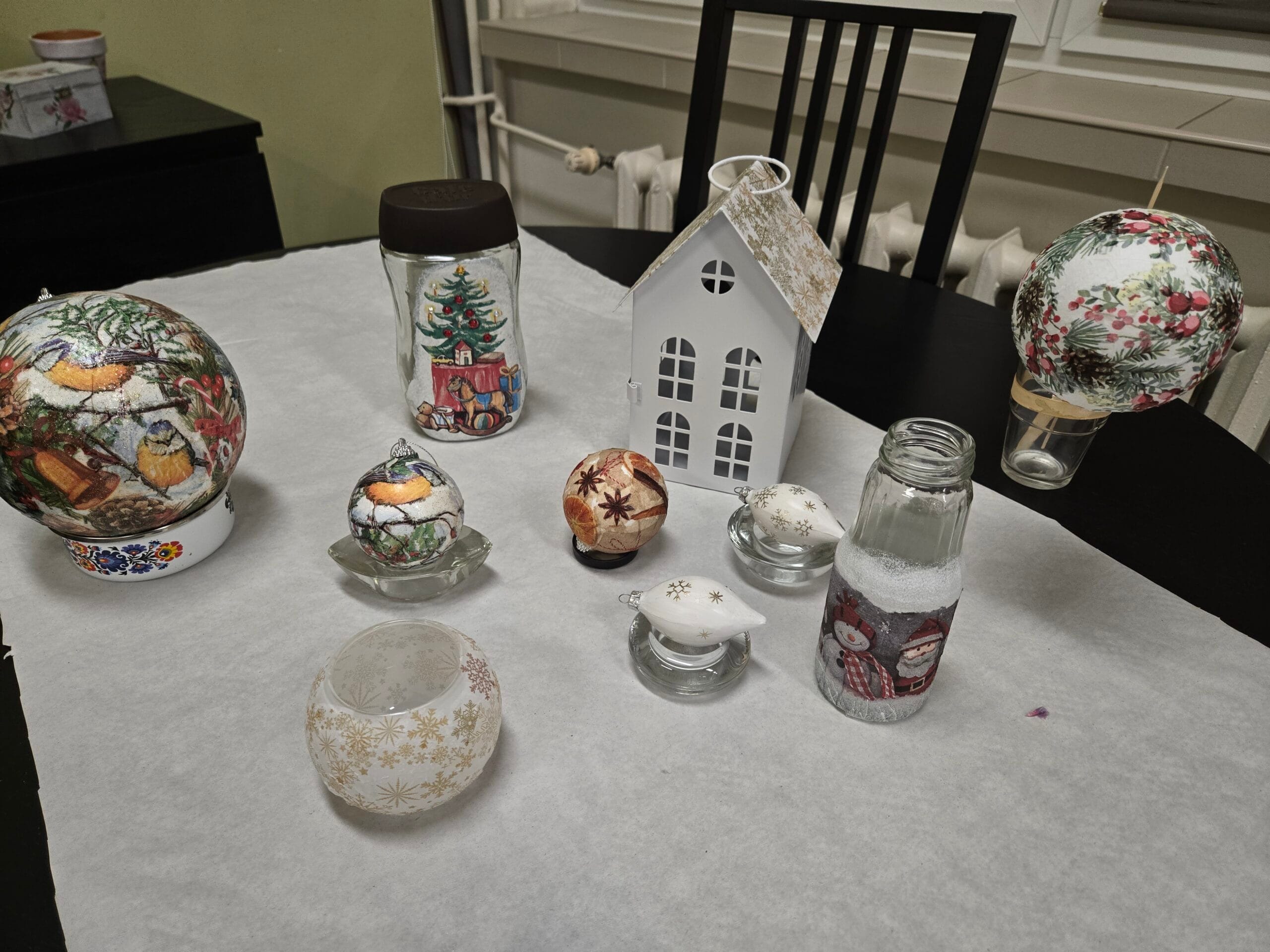 Na stole stoją ozdobione świąteczne bombki, słoiki i butelki oraz biała latarenka w kształcie domku