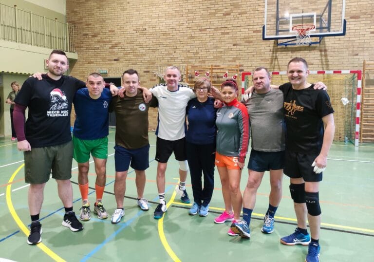 Grupa ośmiu osób w strojach sportowych pozuje do wspólnego zdjęcia na hali gimnastycznej. Stoją w rzędzie, obejmując się ramionami, na tle ceglanej ściany i kosza do koszykówki.