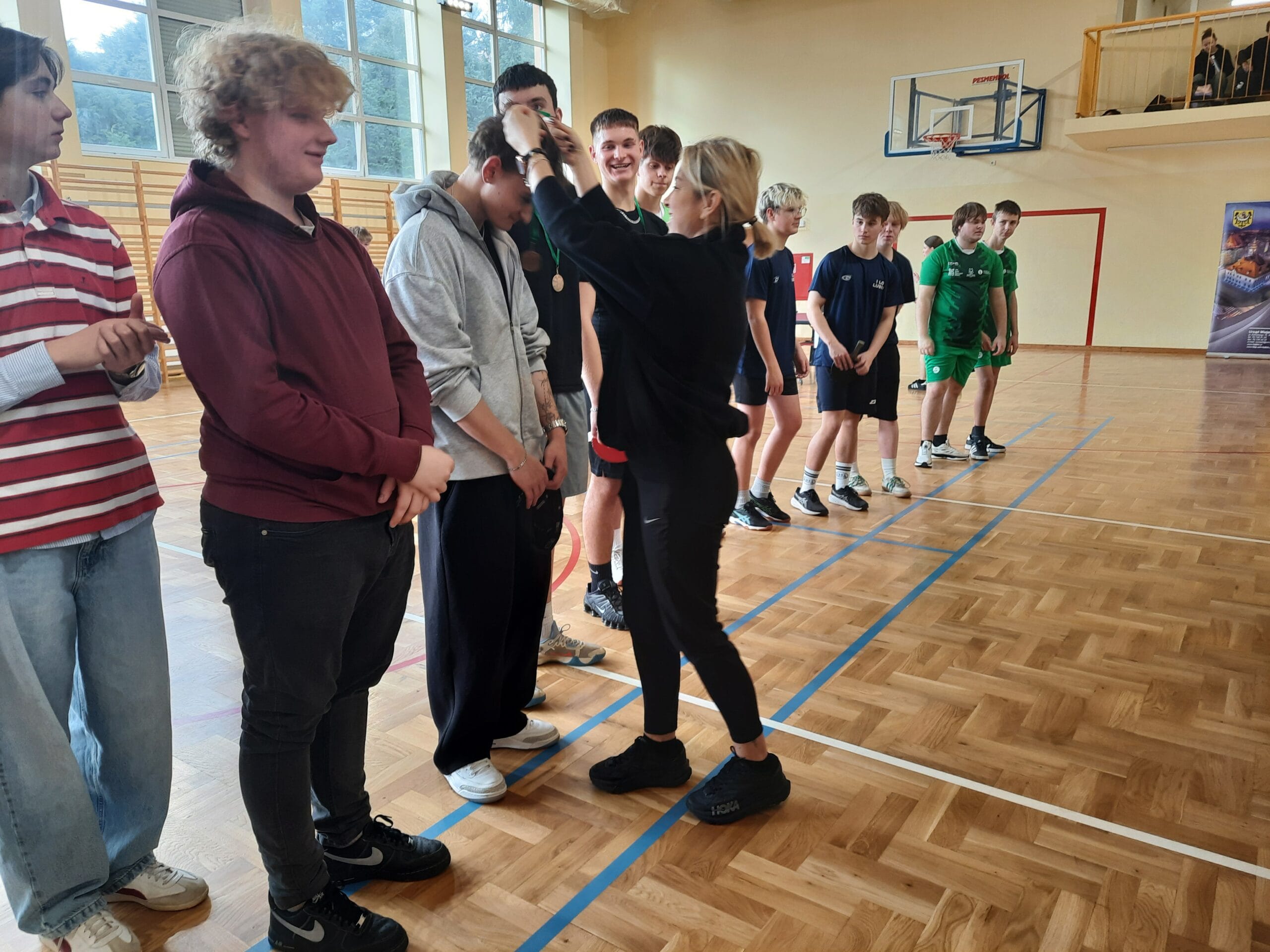 Osoba wręcza medal uczestnikowi stojącemu w rzędzie w sali gimnastycznej. Obok stoją inni uczestnicy w strojach sportowych, część trzyma rakietki do tenisa stołowego. W tle widoczne są duże okna, kosz do koszykówki oraz drewniane drabinki