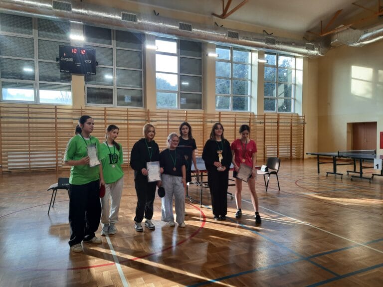 Grupa sześciu osób stoi w sali gimnastycznej przed stołami do tenisa stołowego. Osoby trzymają dyplomy i medale, a jedna z nich puchar. W tle widoczne są duże okna, tablica wyników oraz drewniane drabinki na ścianie