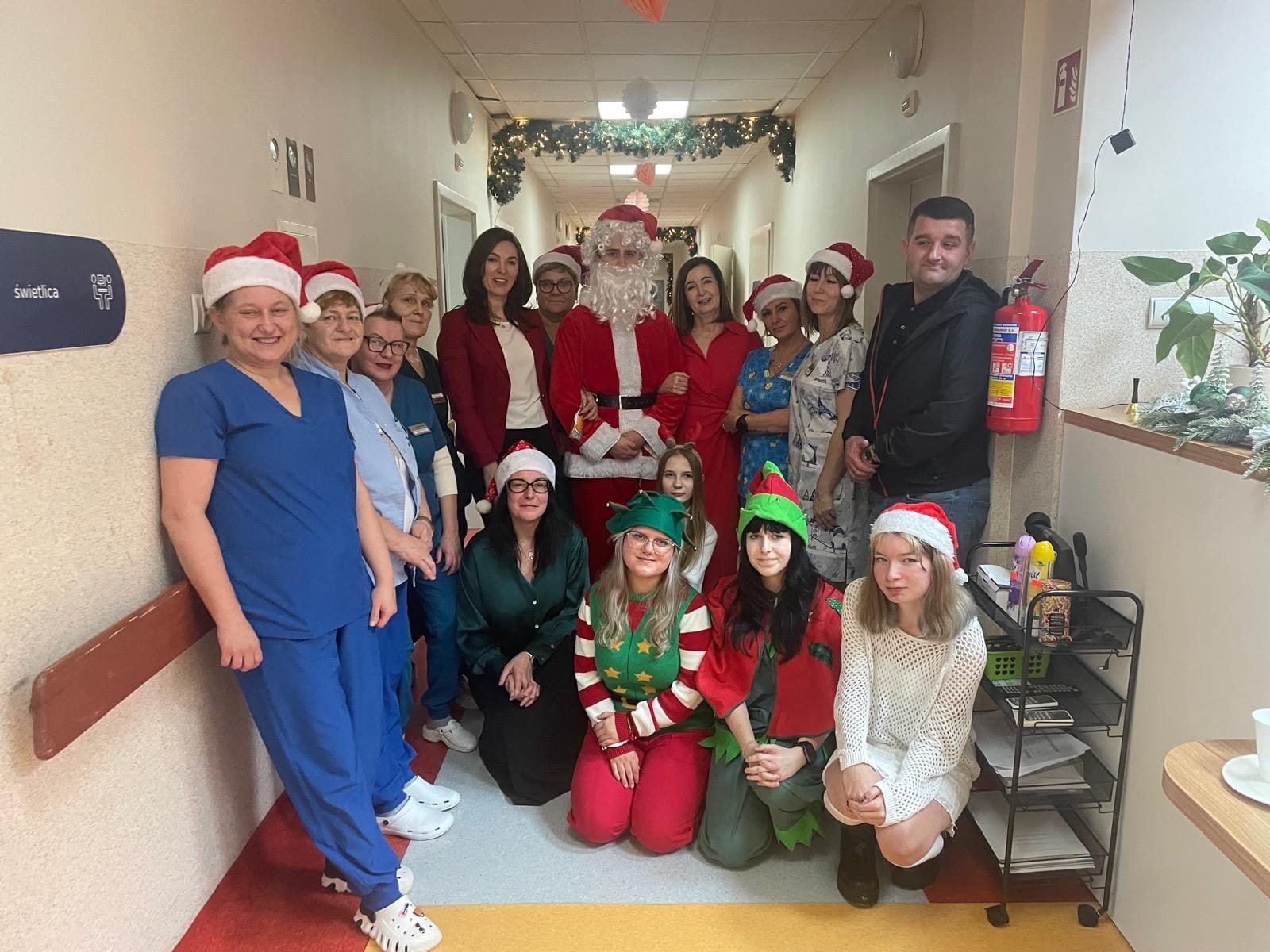Grupa osób w świątecznych strojach, w tym osoba przebrana za Mikołaja, stoi i klęczy w udekorowanym korytarzu. Na ścianach widoczne są girlandy, a w tle ozdoby świąteczne. Kilka osób ma czapki Mikołaja, a dwie osoby stroje elfów. Po prawej stronie znajduje się wózek z rośliną i przedmiotami.
