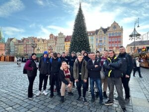 Grupa osób stoi na rynku w otoczeniu kolorowych kamienic, w tle znajduje się duża choinka świąteczna oraz stragany jarmarku bożonarodzeniowego