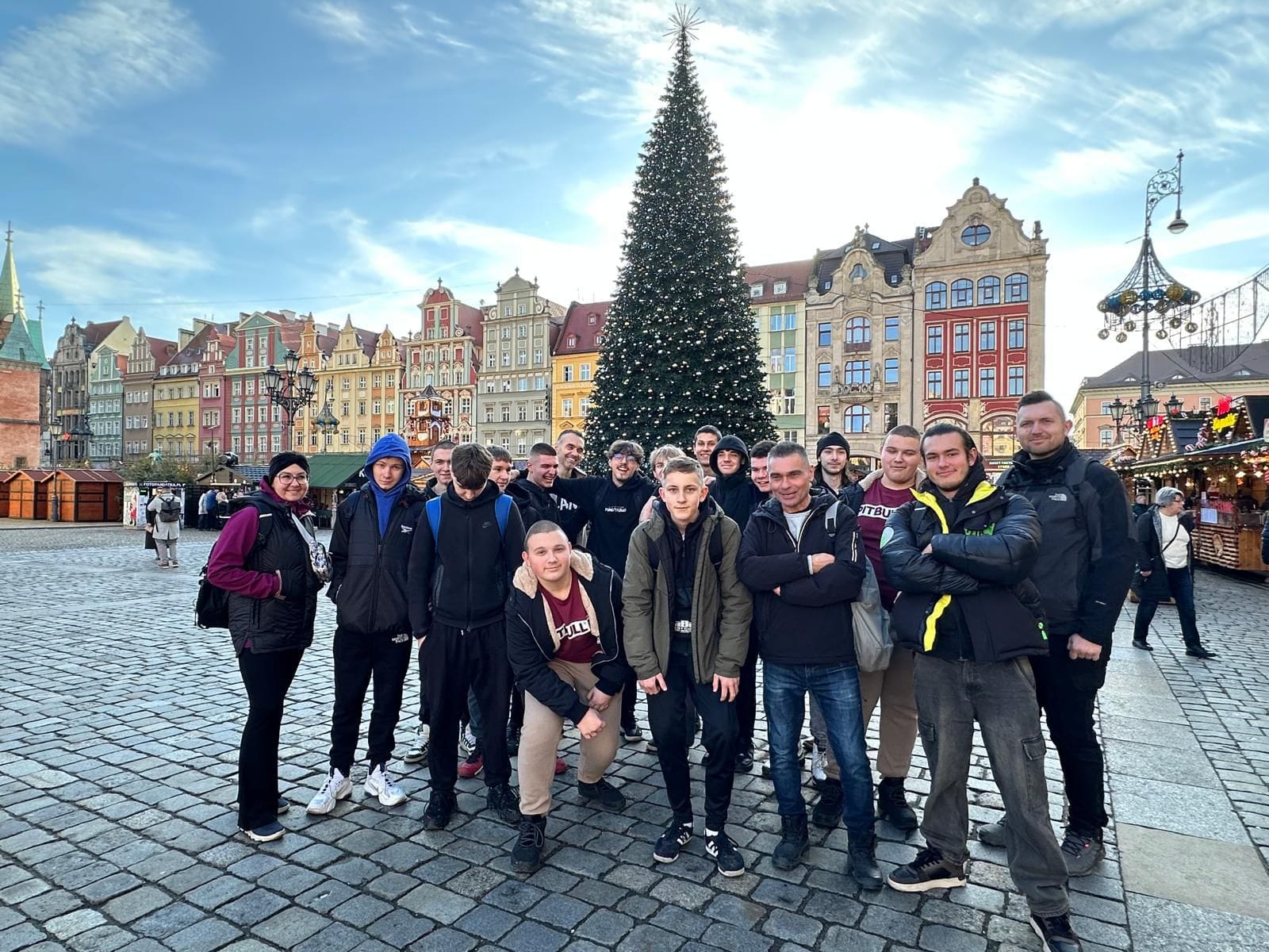 Grupa osób stoi na rynku w otoczeniu kolorowych kamienic, w tle znajduje się duża choinka świąteczna oraz stragany jarmarku bożonarodzeniowego