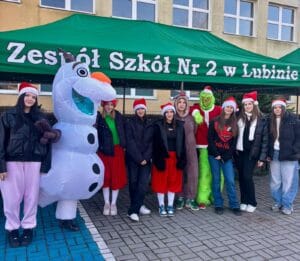 Grupa osób w czapkach Mikołaja stoi przed zielonym namiotem z napisem ‘Zespół Szkół Nr 2 w Lubinie’. Obok nich postać w kostiumie bałwana Olaf oraz osoba w stroju Grincha. W tle budynek szkoły z oknami.