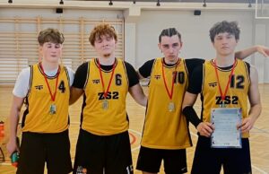 Cztery osoby w żółto-czarnych koszulkach sportowych z numerami 4, 6, 10 i 12 stoją w sali gimnastycznej. Mają na szyi medale, a jedna osoba trzyma dyplom. W tle widoczne są drabinki gimnastyczne i drewniana podłoga