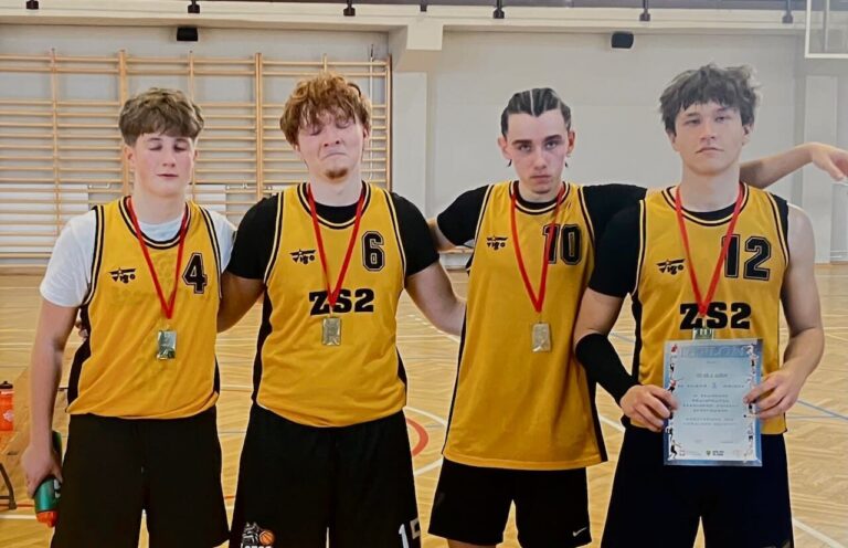 Cztery osoby w żółto-czarnych koszulkach sportowych z numerami 4, 6, 10 i 12 stoją w sali gimnastycznej. Mają na szyi medale, a jedna osoba trzyma dyplom. W tle widoczne są drabinki gimnastyczne i drewniana podłoga