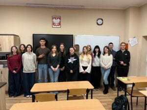 Grupa osób stoi w sali lekcyjnej przed tablicą i ekranem, na ścianie widoczny zegar oraz godło Polski. W tle szafki, a na pierwszym planie kilka pustych drewnianych ławek i krzeseł.