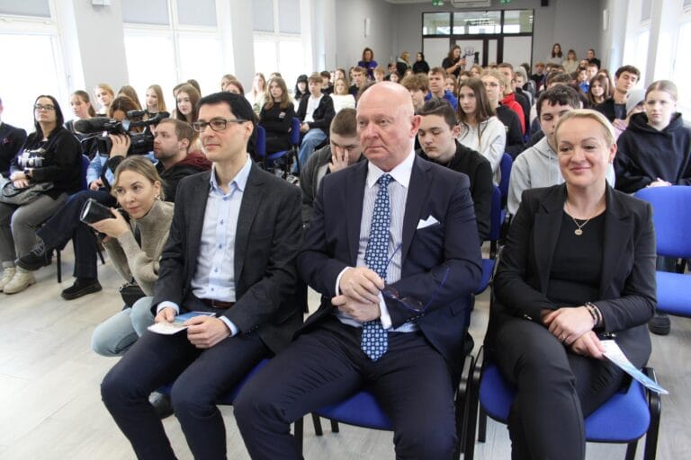 Widok sali konferencyjnej wypełnionej uczestnikami siedzącymi na niebieskich krzesłach. W pierwszym rzędzie trzy osoby w eleganckich strojach trzymają dokumenty. W tle widać dużą grupę osób, część z nich z aparatami i kamerami, a także okna po lewej stronie oraz drzwi wejściowe z tyłu sali