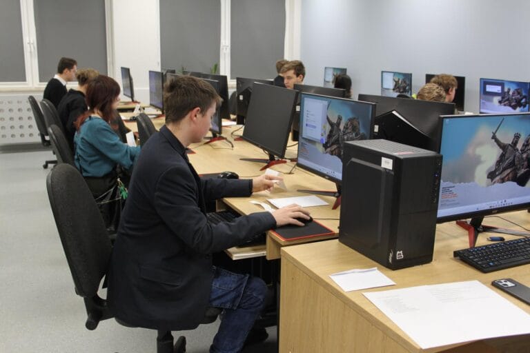 Sala komputerowa z kilkoma stanowiskami. Osoby siedzą przy biurkach, pracując na komputerach stacjonarnych z dużymi monitorami. Na ekranach widoczny jest obraz rycerza w zbroi oraz tekst. Na biurkach znajdują się klawiatury, myszki, podkładki i kartki papieru