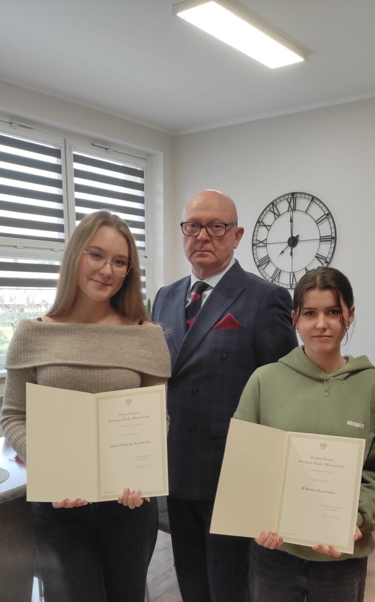 Trzy osoby stoją w pomieszczeniu biurowym, dwie z nich trzymają otwarte dyplomy z widocznym godłem i tekstem. W tle znajduje się okno z poziomymi roletami oraz duży zegar ścienny