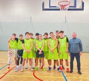 Drużyna koszykarska w zielonych strojach sportowych wraz z opiekunem stoi na sali gimnastycznej pod koszem do gry w koszykówkę.