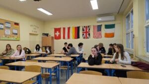 Uczniowie Zespołu Szkół Nr 2 w Lubinie podczas zajęć przygotowawczych do zagranicznych praktyk zawodowych, siedzący w klasie przy ławkach; na ścianie widoczne flagi państw europejskich i USA, symbolizujące międzynarodowy charakter projektu